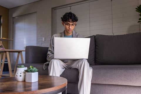 Indian man sitting on gray sofa using laptop for remote work in modern neutral apartment