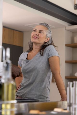 Middle-aged woman leaning on kitchen counter smiling, touching faucet in modern home