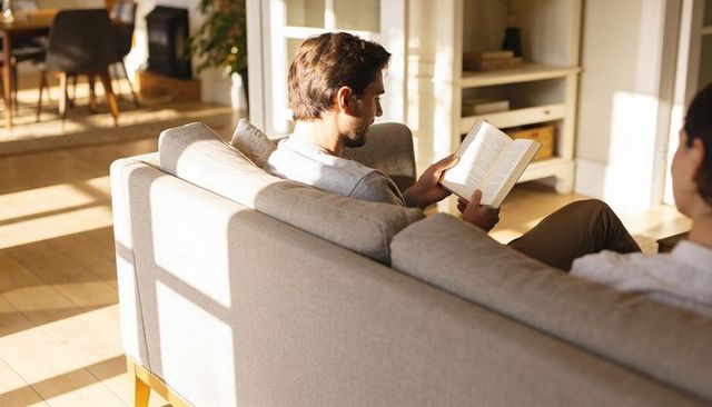 Sunlit living room couple reading on sofa, warm cozy weekend at-home relaxation