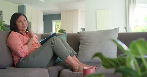 Thoughtful asian woman journaling on sofa in bright contemporary living room for wellness
