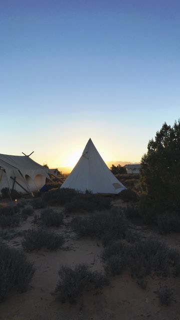 Peaceful scene showing sun setting behind a tipi tent in a desert landscape. Surrounding dry shrubs enhance the desert feel. Suitable for travel advertisements, camping gear promotions, and adventure blogs.