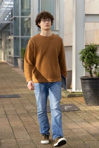 Young man walking on campus holding laptop wearing brown sweater and glasses