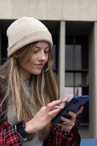 Young woman wearing beanie tapping smartphone while standing by modern concrete building