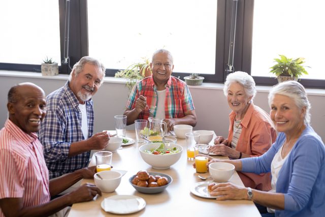 Group of senior friends enjoying breakfast together at a table in a nursing home. They are smiling and appear happy, creating a warm and friendly atmosphere. This image can be used for promoting senior living facilities, healthy eating among the elderly, and community bonding activities.