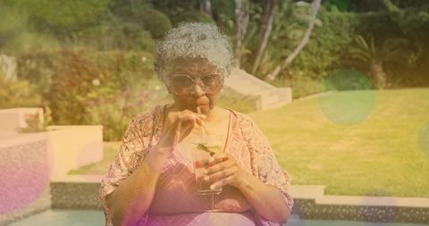 Senior woman sipping citrus cocktail poolside in sunlit backyard while wearing sunglasses
