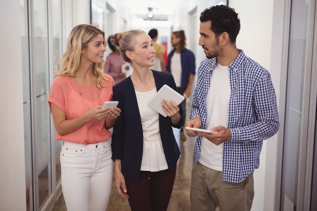 Businesswoman engaging in conversation with creative team members in a modern office corridor. They are holding tablets and smartphones, indicating a tech-savvy work environment. Ideal for use in articles or advertisements about teamwork, modern workplaces, business communication, and professional collaboration.