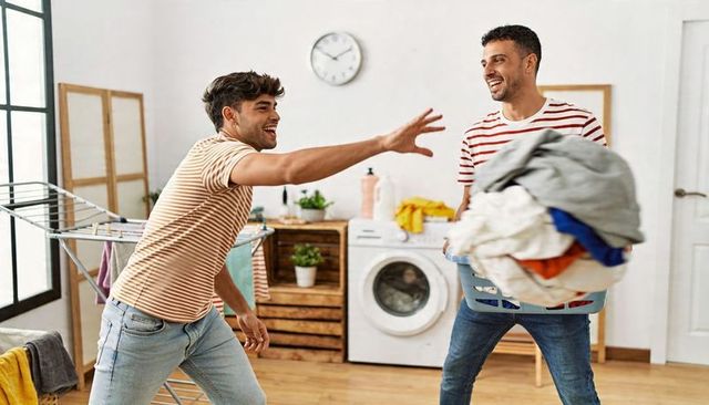 Two friends laughing while carrying laundry basket in bright laundry room, wearing striped shirts