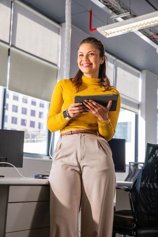 Confident professional standing with tablet in sunlit modern office wearing mustard top