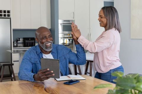 Mature couple celebrating while using tablet and sharing high-five at modern kitchen table