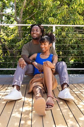 African american couple sitting on wooden deck embracing and smiling under sunlit terrace