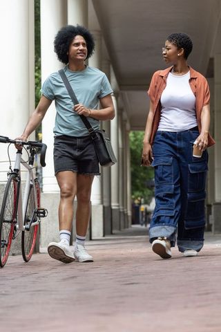 African american friends walking with bicycle and coffee under covered colonnade on campus