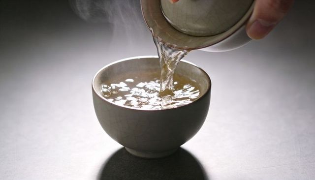 Pouring gaiwan tea into crackle-glazed ceramic cup with rising steam, minimal tabletop