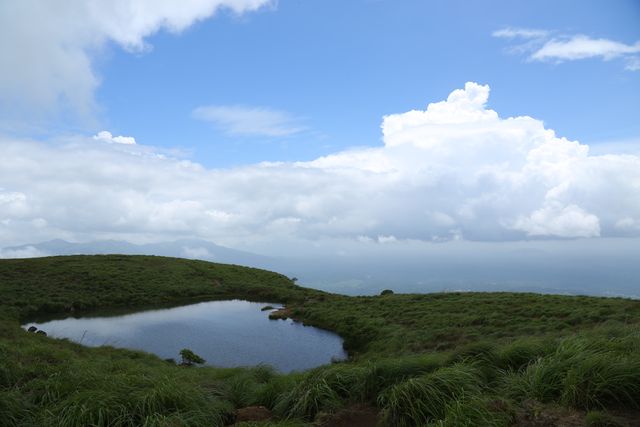 This picturesque scene captures a serene hilltop pond surrounded by lush greenery under a partly cloudy sky. Ideal for promoting nature tourism, hiking spots, outdoor activities, and environmental campaigns. It can also be used in travel magazines, blogs, and posters promoting tranquility and natural beauty.