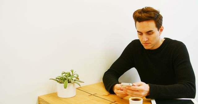 Young man sitting in a minimalist cafe, using his smartphone. Small white plant pot and a coffee cup are on the wooden table. Man appears focused on phone screen. Ideal for illustrating concepts of modern lifestyle, technology use, cafe culture, or casual settings.