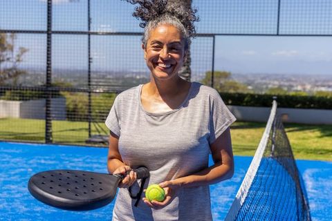 African american mature woman smiling on outdoor padel court holding racket and ball