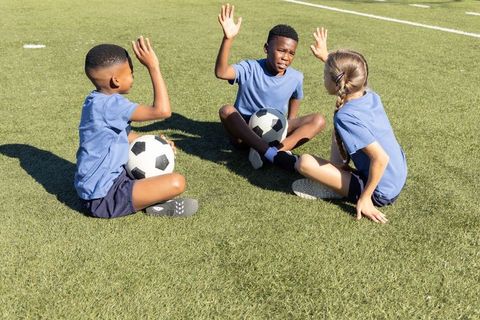 Diverse youth soccer players sitting on turf raising hands during sunny practice