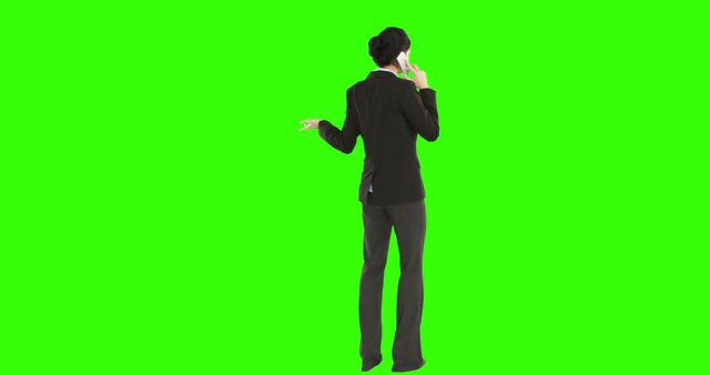 A young Caucasian businesswoman is engaged in a phone conversation while gesturing with her other hand, against a green screen background with copy space. Her professional attire and active stance suggest a dynamic work-related discussion.