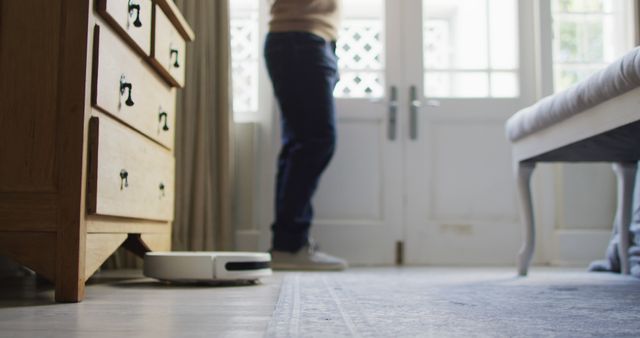 Person standing in bedroom with a robotic vacuum cleaner in operation, depicting modern home automation and convenience. Useful for illustrating smart home technology, daily cleaning routines, and modern living environments.