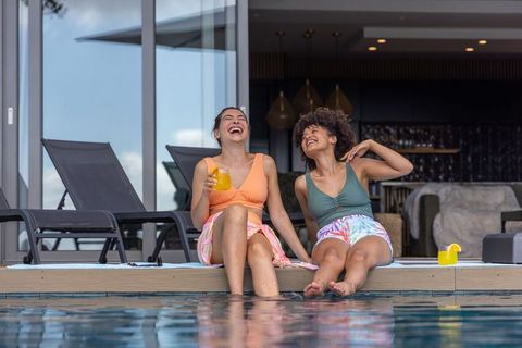 Diverse friends laughing poolside drinking orange cocktails on resort deck enjoying summer