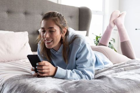 Young woman relaxing on bed scrolling smartphone in cozy pastel bedroom