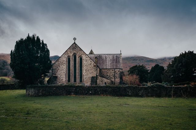 This image depicts a historic stone church set amidst a scenic mountain landscape under a cloudy sky. The building showcases ancient architectural style typical of rural settings. Ideal for use in projects related to history, religion, travel, or rural life.