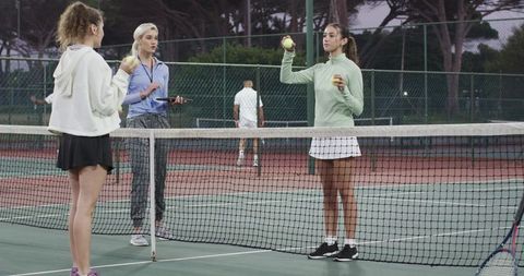 Female coach coaching two players holding tennis balls with tablet on outdoor court at dusk
