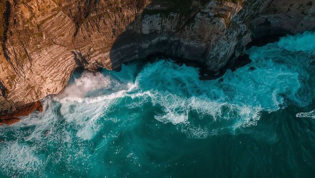 Turquoise waves crashing against layered sandstone cliffs creating foam, sea cave spray