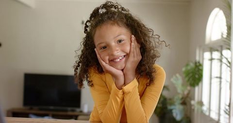 Smiling girl leaning chin on hands at home wearing mustard top, cozy daylight family moment