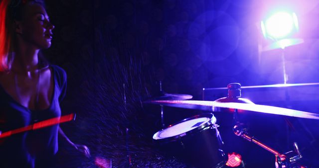 Female musician playing drums on a stage with vibrant, dramatic lighting creating a blue and purple glow. Useful for promoting music events, concerts, live performances, and vibrant nightlife scenes. Also suitable for illustrating music-related articles, blogs, and advertisements.