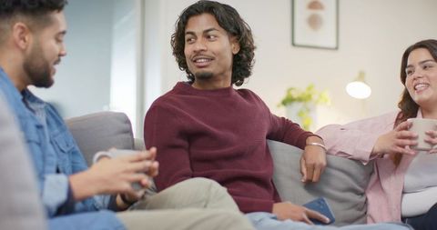 Friends relaxing on couch in cozy living room holding mug and smartphone, smiling and chatting