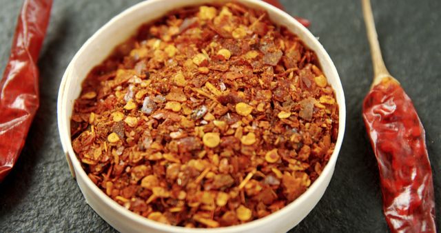 A bowl of crushed red chili flakes is presented on a dark slate surface, flanked by whole dried chilies, with copy space. Red chili flakes are a versatile spice used to add heat and flavor to a variety of dishes across different cuisines.