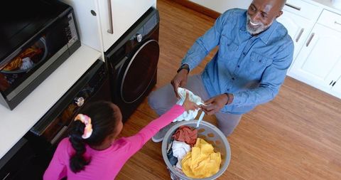 African american father kneeling doing laundry with daughter handing clothes, smiling bond