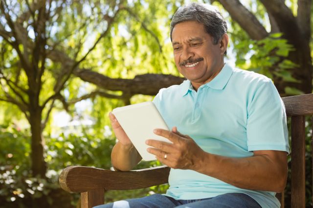 Senior man sitting on a wooden bench in a garden, using a digital tablet. He is smiling and appears relaxed, enjoying the outdoor setting. Ideal for use in articles or advertisements related to senior lifestyle, technology use among the elderly, outdoor activities, or digital literacy.