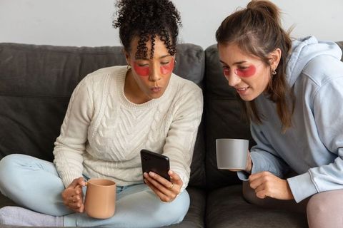 Diverse friends relaxing on couch with under-eye patches and coffee mugs in cozy knitwear