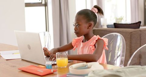 School-age girl studying on laptop at dining table with orange juice and headphones