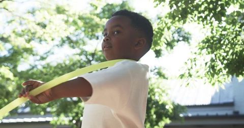 Sunlit boy playing with yellow-green ribbon under leafy canopy during outdoor summer play