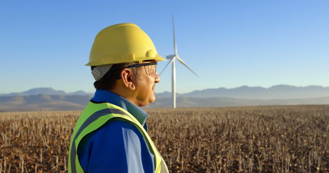 Engineer wearing helmet and safety vest at wind farm with wind turbines in background. He is looking at the wind turbine against mountain backdrop. Useful for themes relating to renewable energy, sustainability, engineering, professional work, and environmental conservation.
