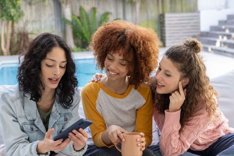Diverse women relaxing poolside and sharing smartphone moments with coffee