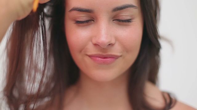 Close-up depicts a woman smiling with her eyes closed while her hair is being combed against white backdrop. Great for illustrating beauty routines, self-care, well-being advertising, and tutorials on hair care.