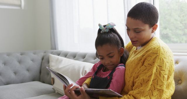 Mother and daughter share a special moment reading a storybook on a gray couch in a cozy living room. Ideal for themes of family bonding, parent-child relationships, quality time, education, and home life. Perfect for use in family-oriented content, educational materials, parenting blogs, and social media posts about childhood and nurturing.