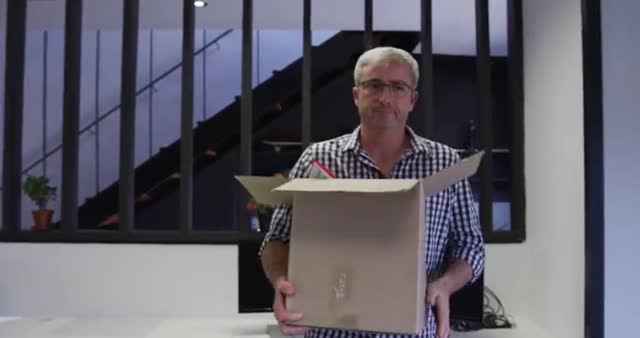 Mature Caucasian male with grey hair and glasses is moving offices, carrying a cardboard box to his new workspace. He wears a checked shirt and stands in a modern office with a staircase in the background. This can be used to depict scenes related to job transitions, moving offices, or general corporate and business concepts.