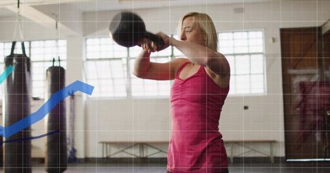 Woman swinging kettlebell in gym wearing pink tank top during strength and cardio training