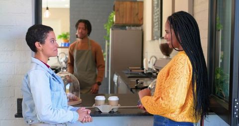 Two women chatting with african american barista at modern cafe counter with takeaway cups