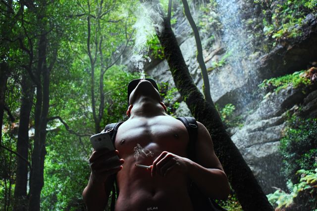 Man enjoying a moment of relaxation while smoking a cigarette in lush forest. Tall trees and greenery surrounding him create perfect wilderness setting. It can be used in themes of outdoor lifestyles, tranquility in nature, and human connection with environment.