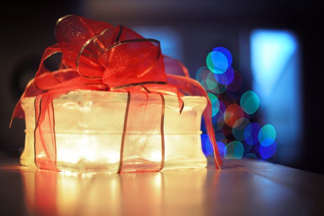 Illuminated gift box wrapped with red ribbon, glowing warmly on a table with colorful, blurred bokeh lights from a Christmas tree in the background. Ideal for holiday greetings, festive advertisements, or decorating ideas promoting seasonal themes.