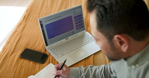 Asian Man Taking Notes with Laptop on Bed