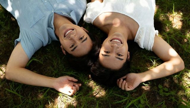 Young couple lying head-to-head on grass smiling under sun-dappled trees in park