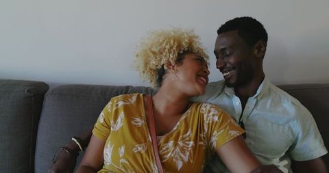 Diverse couple cuddling on grey sofa in cozy living room, woman leaning and smiling