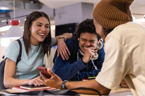 Laughing diverse students sharing smartphone while studying together at campus lounge