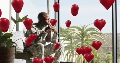 Contemplative soldier sitting by window surrounded by floating red heart balloons
